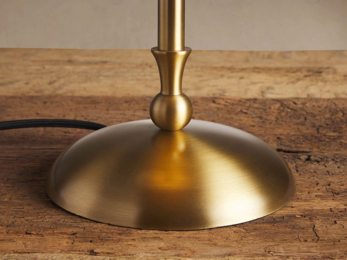 Archard Table Lamp with a brass finish, domed shade, slender stem, and round base, standing on a wooden table against a plain wall.