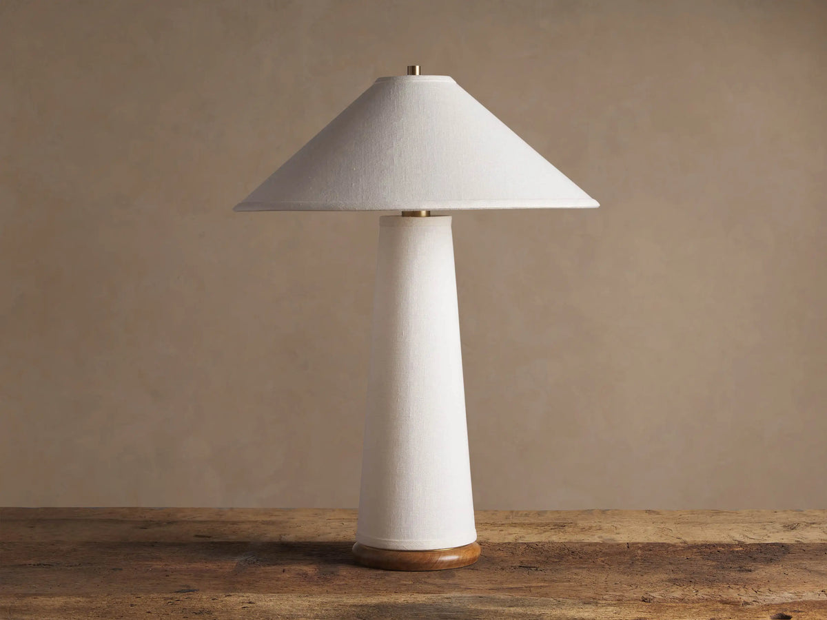 Ombra Table Lamp with a wide, conical light linen shade & tapered body, teak wood base, & brass accents, on a wooden table.