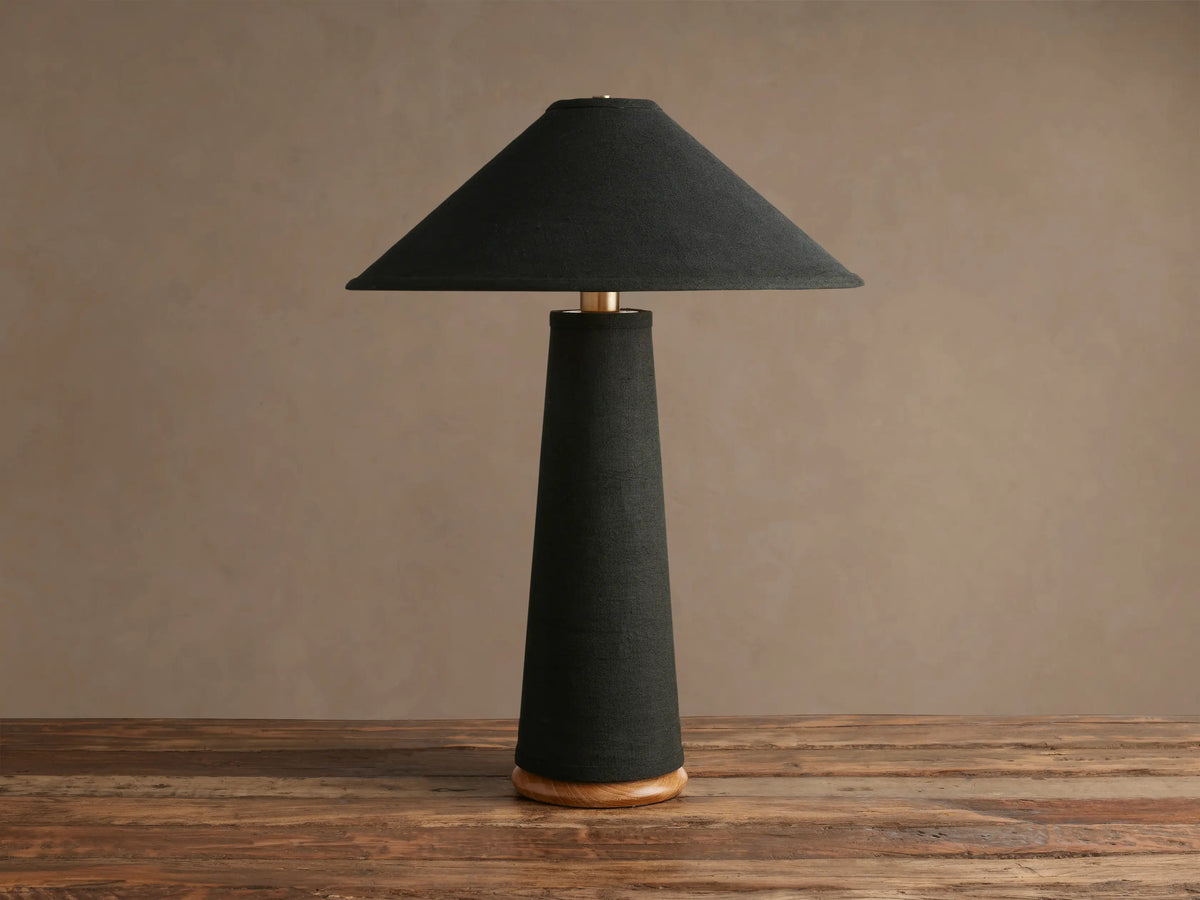 Ombra Table Lamp with black conical shade and tapering black fabric stem, brass accent, and teak wood base, on a wooden table.