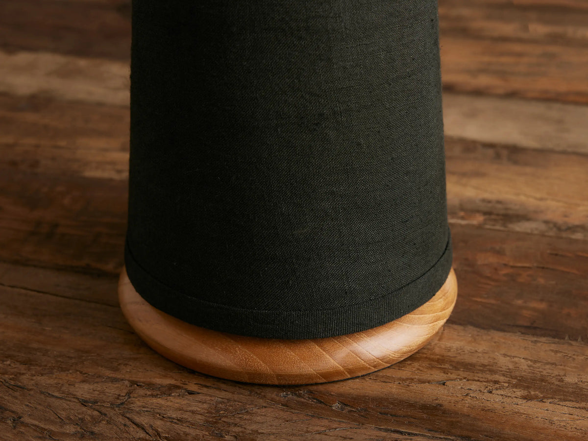 Close-up of Ombra Table Lamp base: cylindrical black linen shade sits on a rounded light teak wood base on a dark wooden surface.