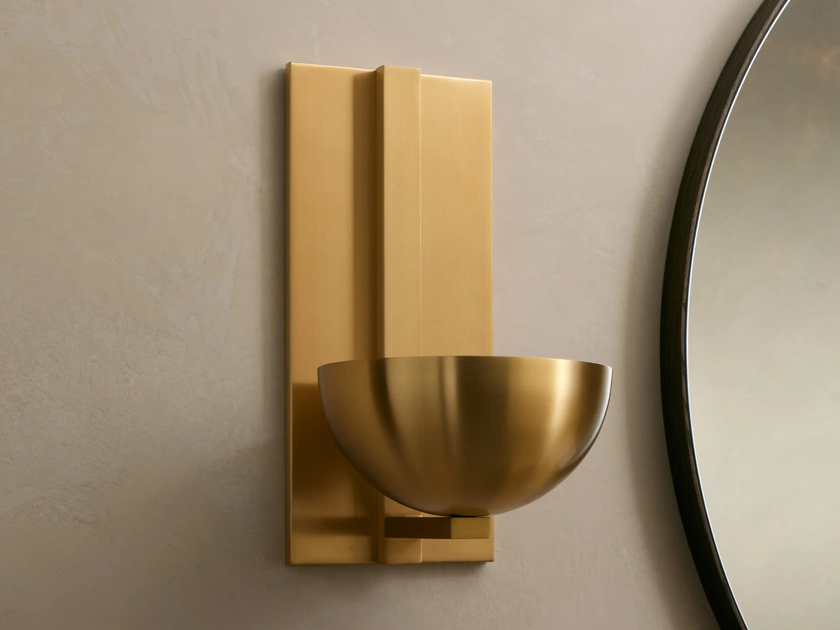 Belmira Sconce, brass finish, wall-mounted, with a rectangular backplate and bowl shade, casting an upward glow beside a mirror.