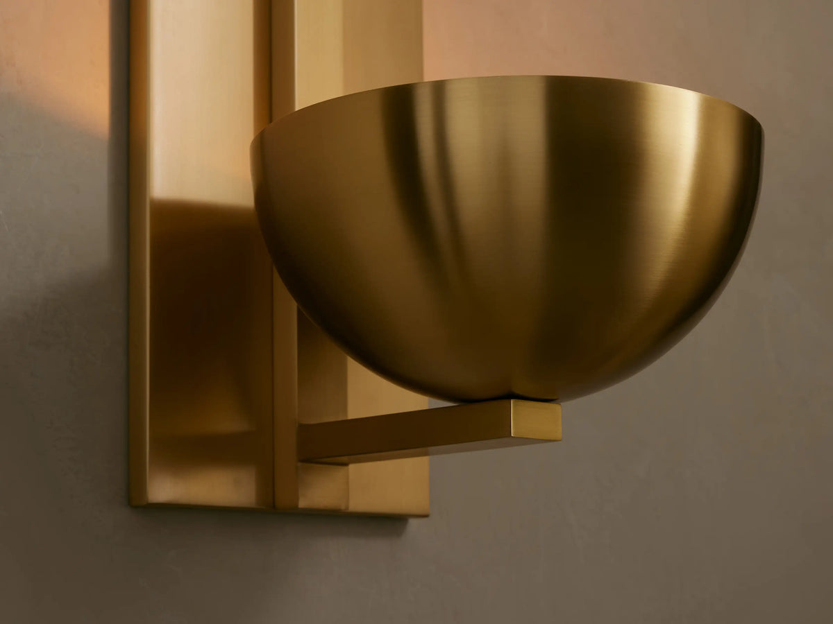 Vertical brass Belmira Sconce with rectangular backplate and bowl-style shade on a textured wall, partial mirror right.