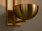 Vertical brass Belmira Sconce with rectangular backplate and bowl-style shade on a textured wall, partial mirror right.