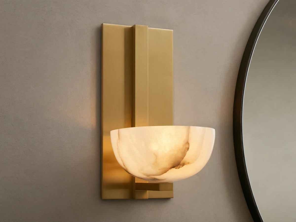 Belmira Sconce: Brass-finished wall light with glowing, bowl-shaped alabaster shade on a textured wall next to a mirror.