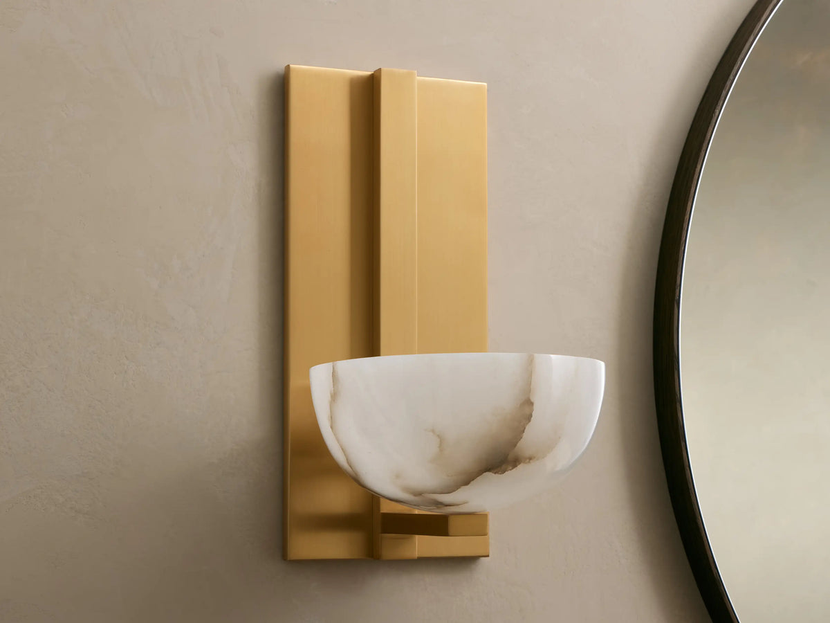 Belmira Sconce: Brass-finished wall light with glowing, bowl-shaped alabaster shade on a textured wall next to a mirror.