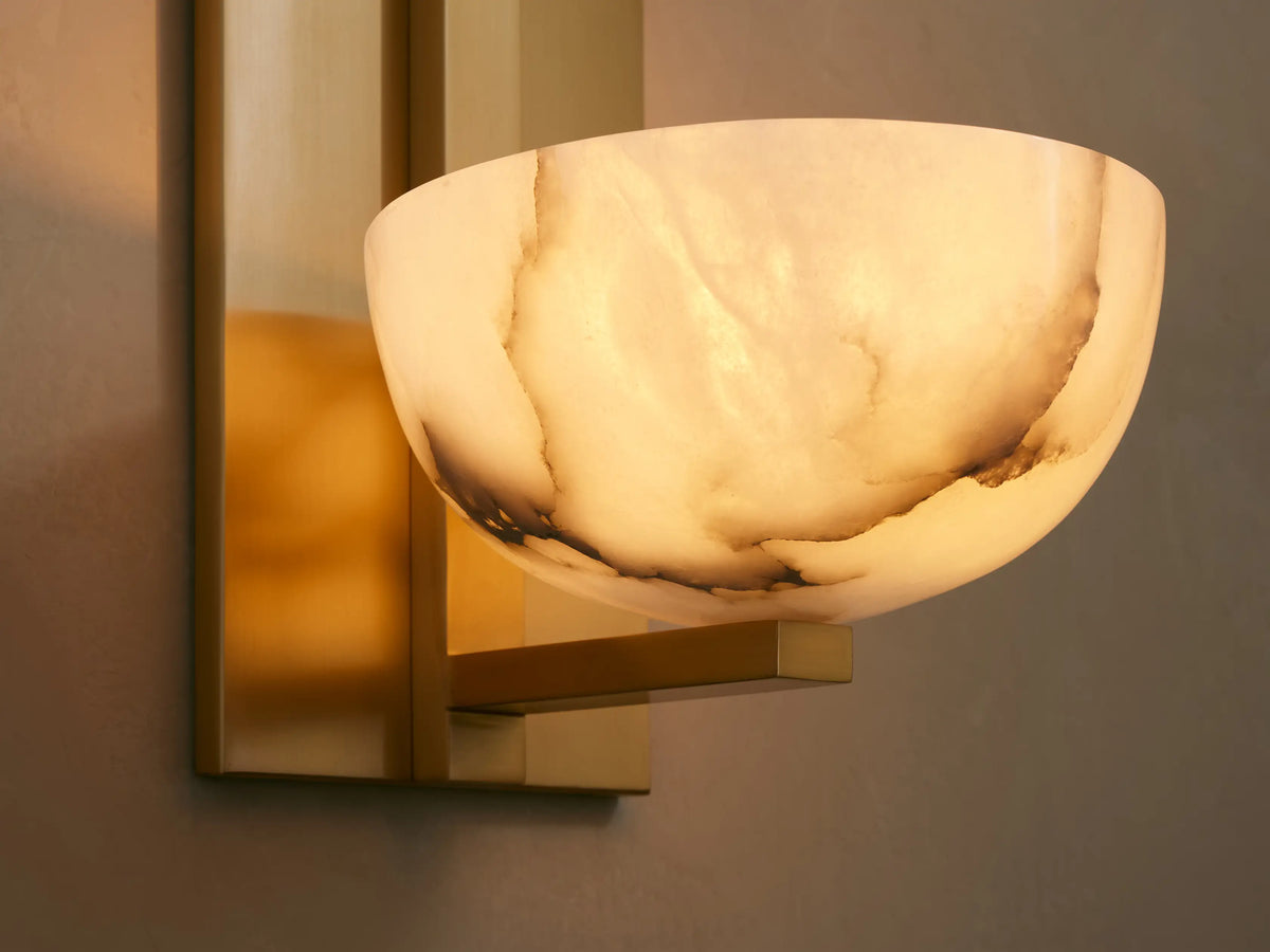 Belmira Sconce: Brass-finished iron wall sconce with a white alabaster bowl shade featuring natural brown veining on a tan wall.
