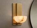 Belmira Sconce: Brass-finished wall light with glowing, bowl-shaped alabaster shade on a textured wall next to a mirror.