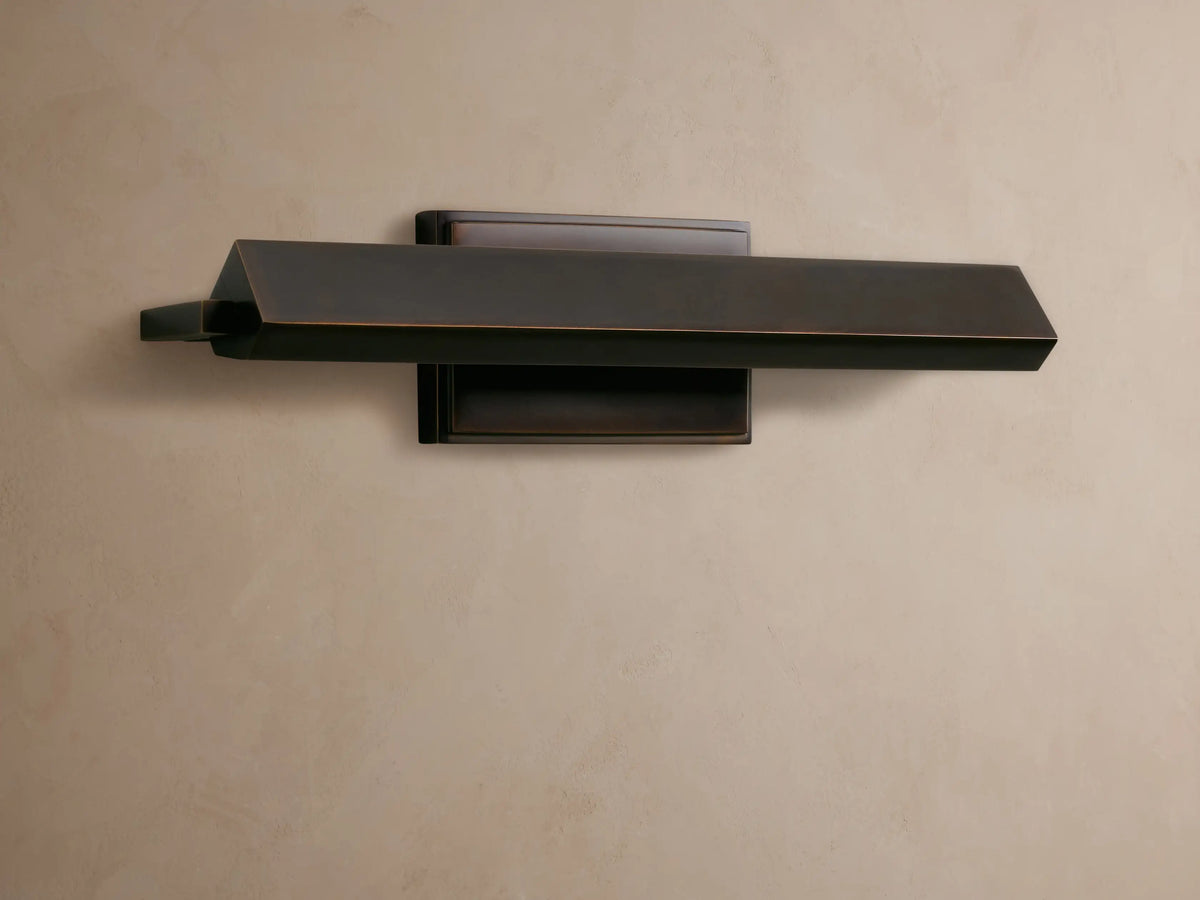 Zaeden Picture Light, dark bronze-plated with a horizontal angled bar and rectangular backplate, mounted on a textured brown wall.