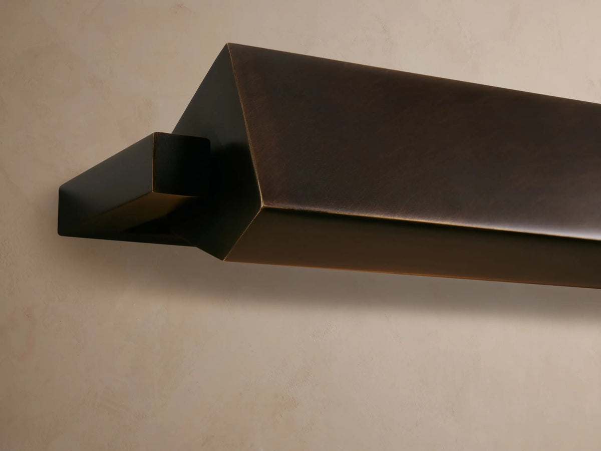 Zaeden Picture Light, an angular dark bronze fixture, mounted on a textured wall, emitting a warm golden glow downwards.