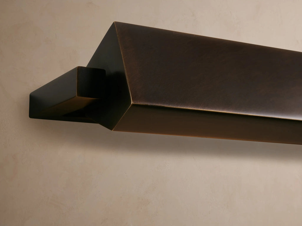 Zaeden Picture Light, an angular dark bronze fixture, mounted on a textured wall, emitting a warm golden glow downwards.