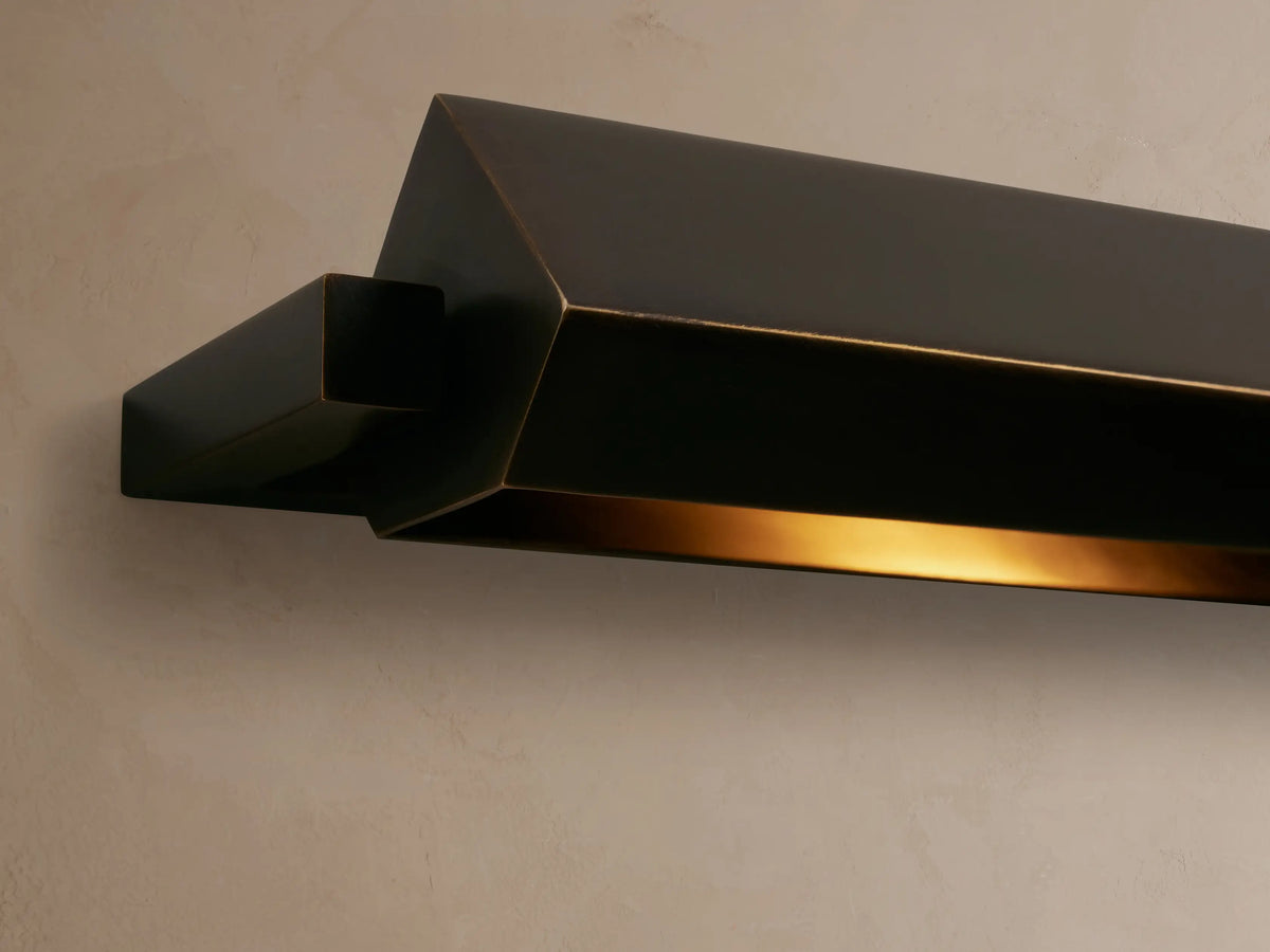 Close-up of Zaeden Picture Light, an angular bronze-plated fixture mounted on a wall, emitting warm golden light.