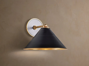 Arno Sconce: matte black conical shade with polished brass interior & trim, brass arm & circular backplate on brown wall, lit.