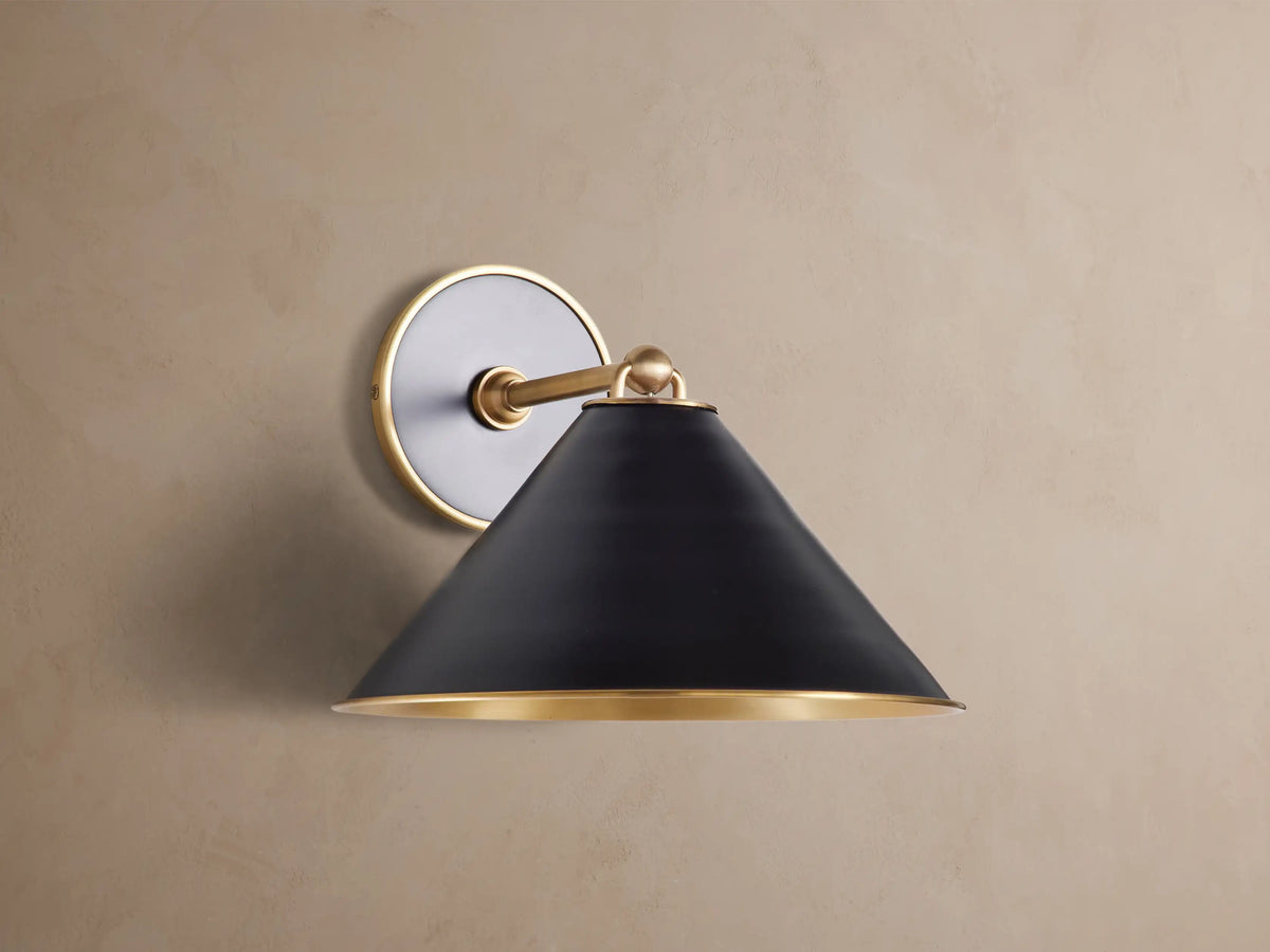 Arno Sconce: matte black conical shade with polished brass interior & trim, brass arm & circular backplate on brown wall, lit.