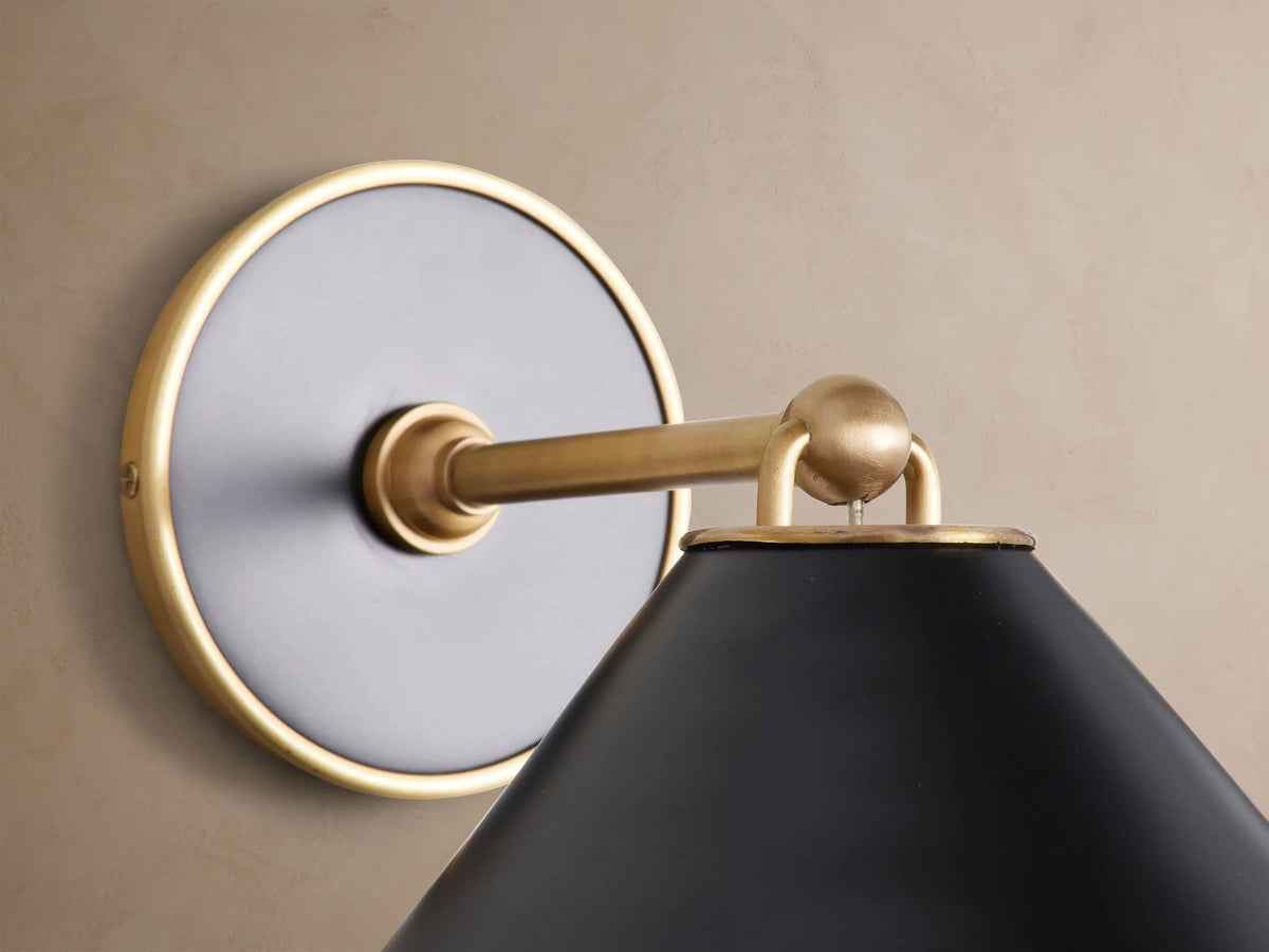 Arno Sconce with a matte black conical shade and polished brass interior, mounted on a textured neutral wall.