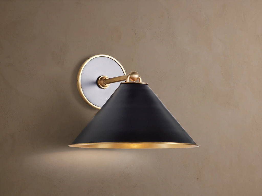 Arno Sconce: matte black conical shade with polished brass interior & trim, brass arm & circular backplate on brown wall, lit.