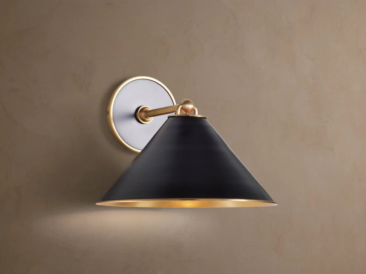 Arno Sconce: matte black conical shade with polished brass interior & trim, brass arm & circular backplate on brown wall, lit.