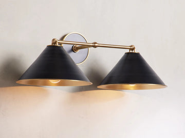 Arno Double Sconce with matte black cone-shaped shades, brass arm, polished brass interior, and round backplate on a wall.