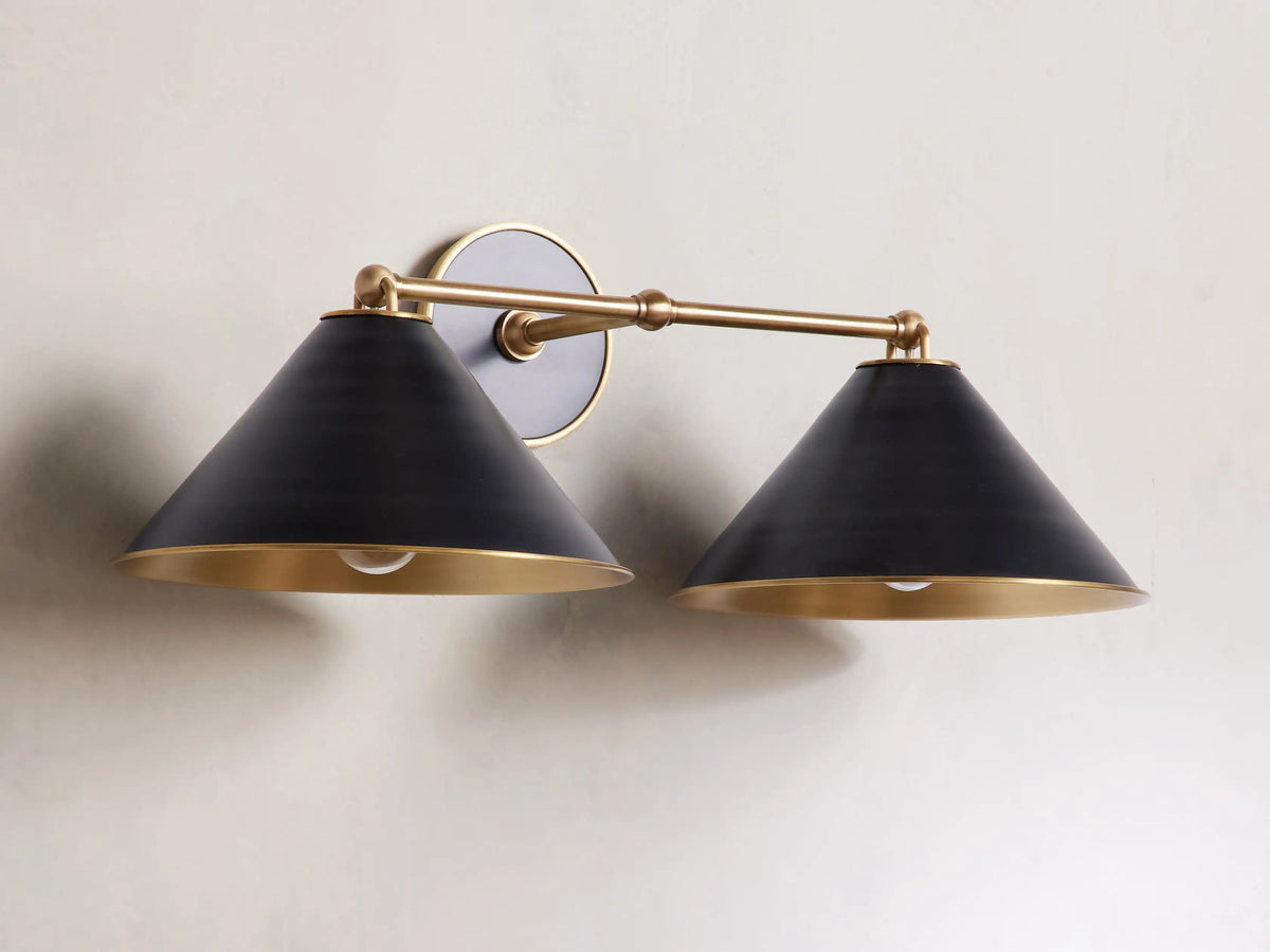 Arno Double Sconce with matte black cone-shaped shades, brass arm, polished brass interior, and round backplate on a wall.