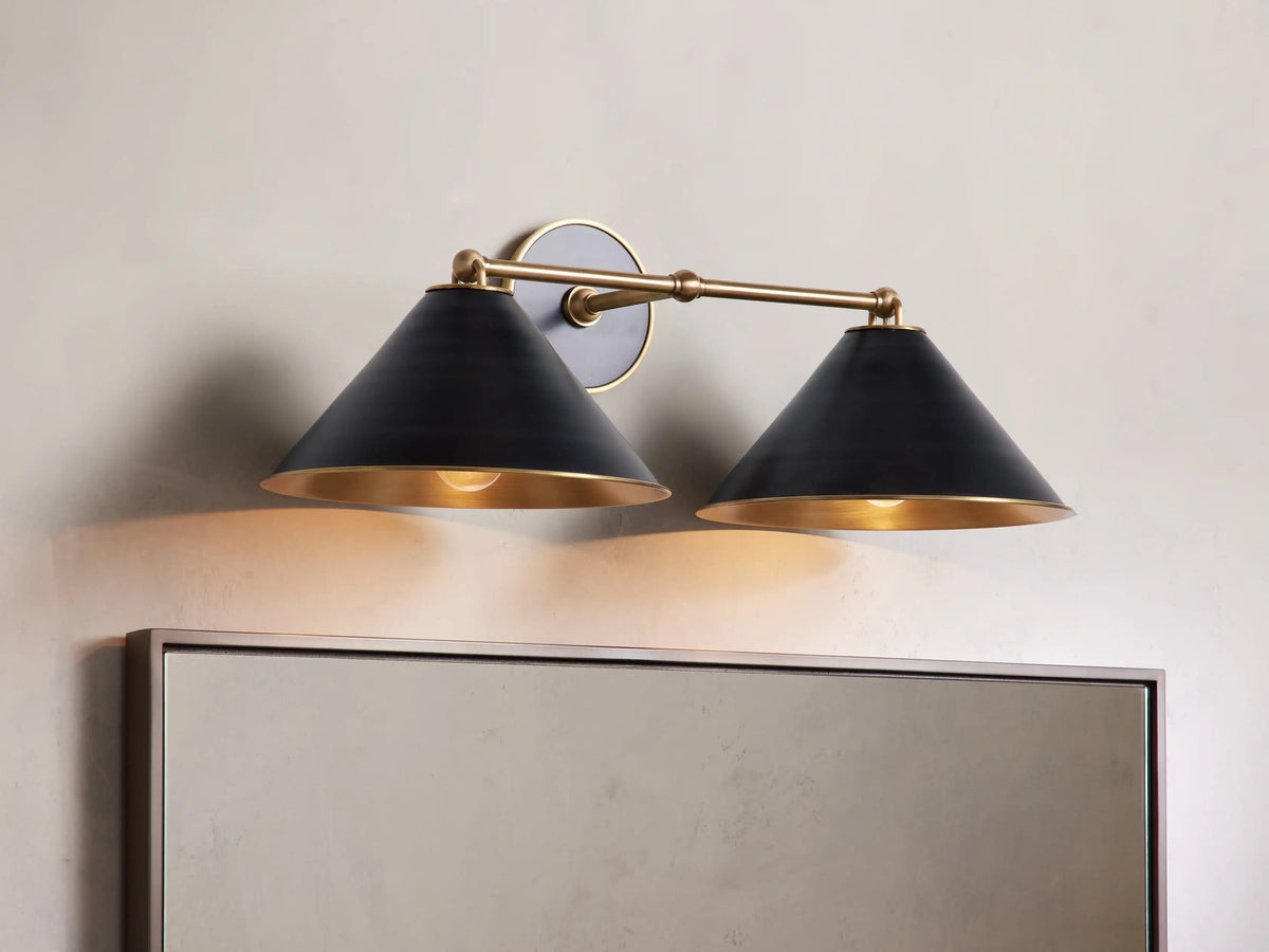 Black Arno Double Sconce with polished brass interior, mounted on wall, casting warm light. Accompanied by a matching chandelier.