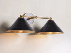 Arno Double Sconce with matte black cone-shaped shades, brass arm, polished brass interior, and round backplate on a wall.