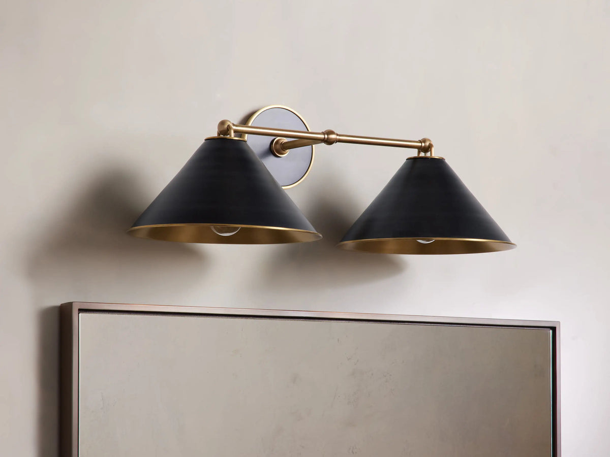 Arno Double Sconce with black conical shades, brass interiors, mounted on a wall above a mirror, casting warm light.