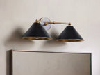 Arno Double Sconce with black conical shades, brass interiors, mounted on a wall above a mirror, casting warm light.
