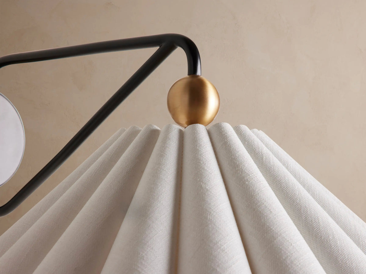 Haylen Sconce: Black iron wall light with antique brass accents, featuring a pleated, light-colored fabric cone shade, against a tan wall.