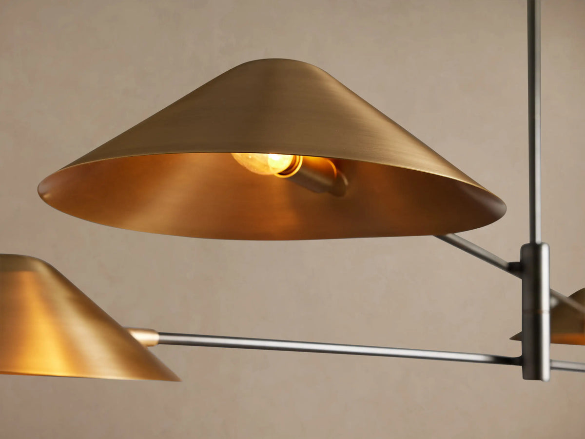 Simms Chandelier: Four conical brass shades pointing downwards on dark metal pivoting arms, hanging centrally against a neutral wall.