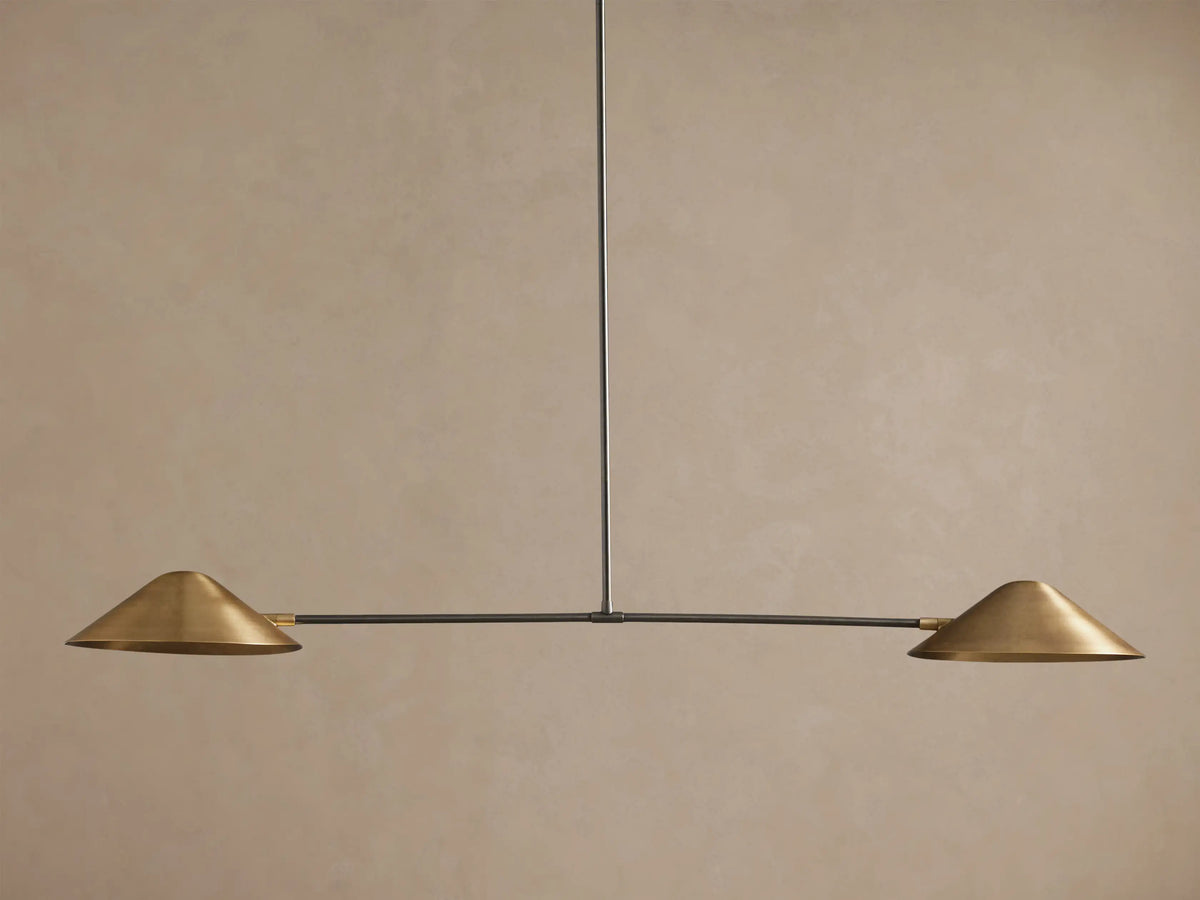 Simms Linear Chandelier with two downward-facing brass conical shades on a dark horizontal bar, suspended against a taupe wall.
