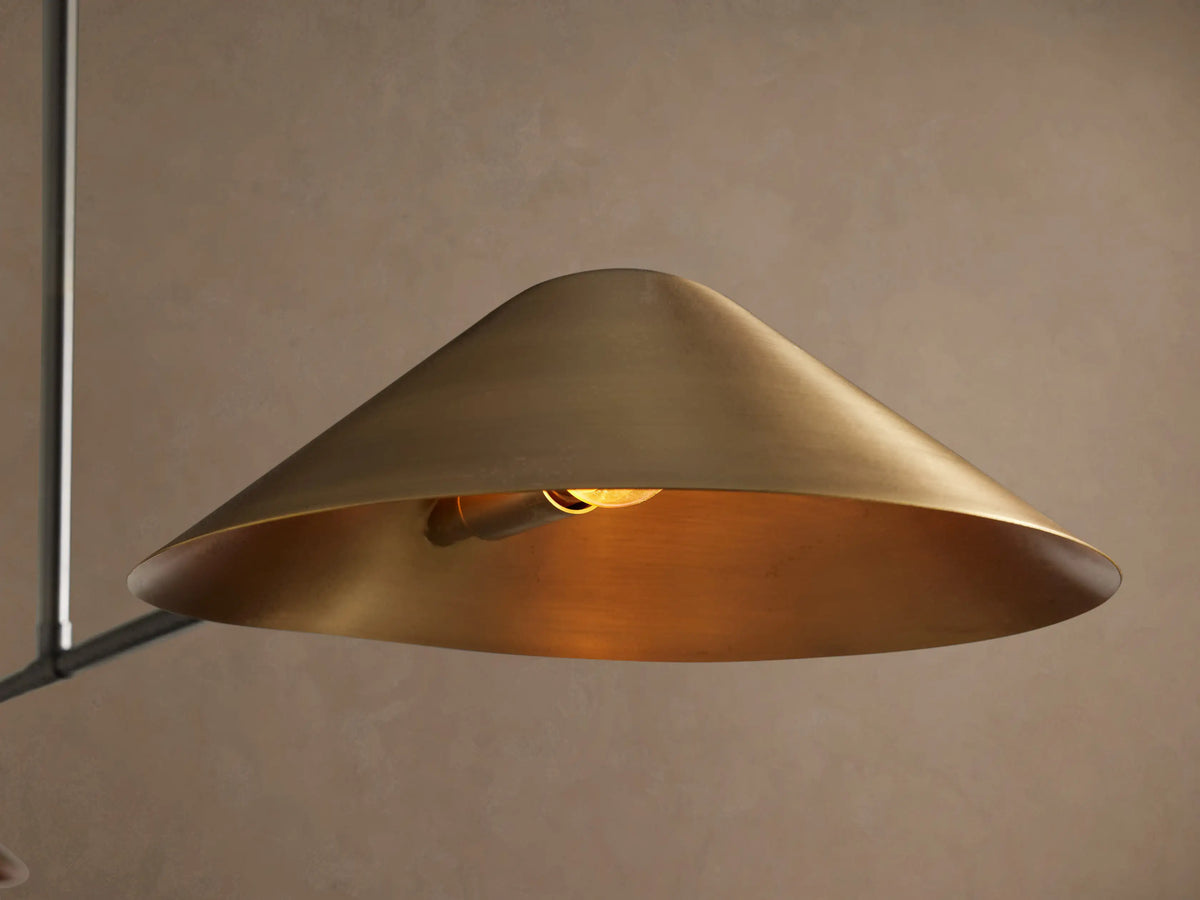 Simms Linear Chandelier with two conical antiqued brass shades on a dark linear bar, suspended by a matching rod against a neutral wall.