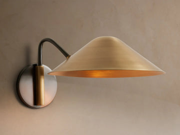 Simms Sconce: A large, conical polished brass shade emits a warm glow, attached to a dark curved arm & round backplate.