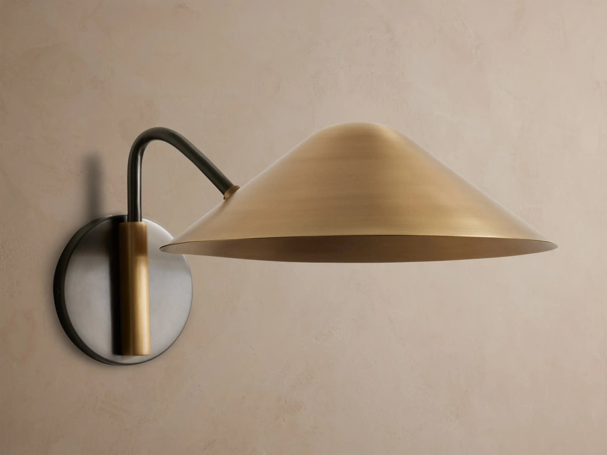 Simms Sconce: A large, conical polished brass shade emits a warm glow, attached to a dark curved arm & round backplate.