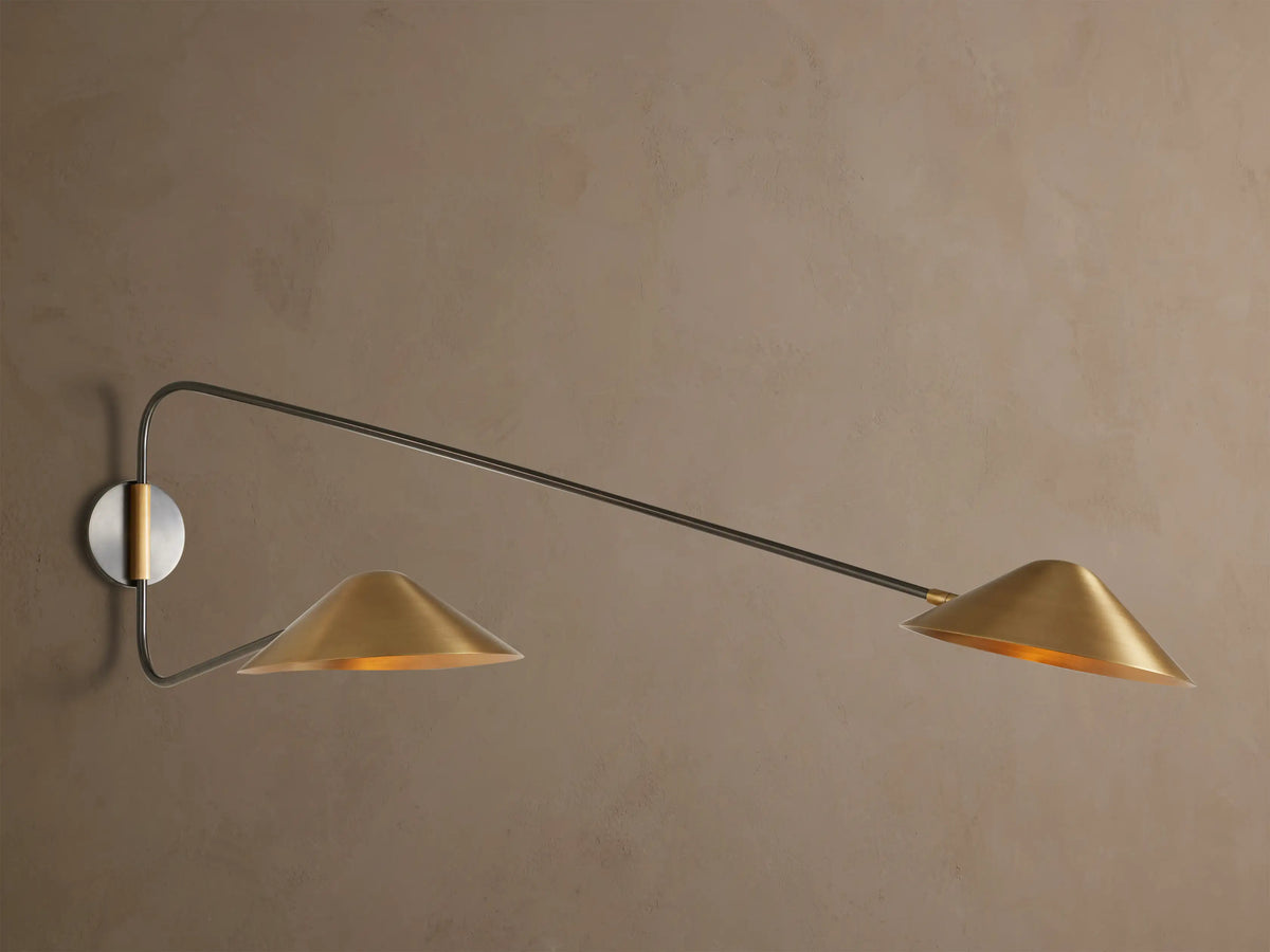 Simms Long Arm Double Sconce: Two antiqued brass conical lights with dark metal arms extending from a silver-toned backplate on a wall.
