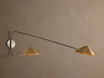 Simms Long Arm Double Sconce: Two antiqued brass conical lights with dark metal arms extending from a silver-toned backplate on a wall.