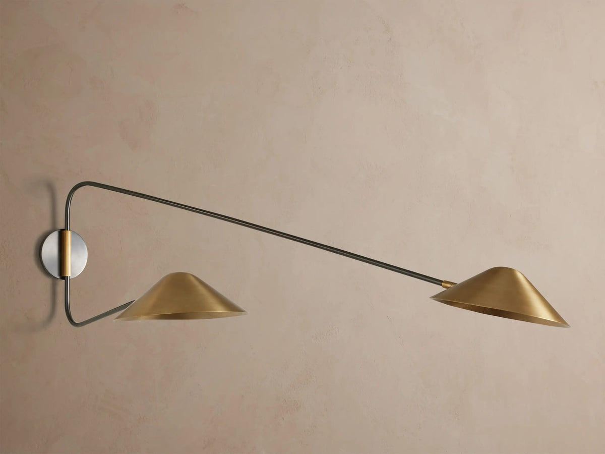 Simms Long Arm Double Sconce: Two antiqued brass conical lights with dark metal arms extending from a silver-toned backplate on a wall.