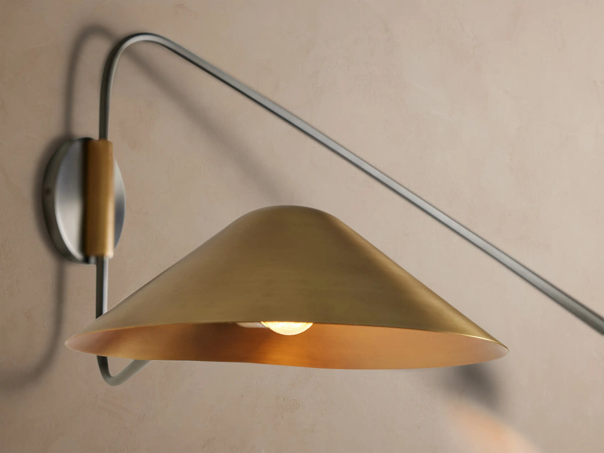 Simms Long Arm Double Sconce: dark curved metal structure with two polished brass conical shades against a beige wall.