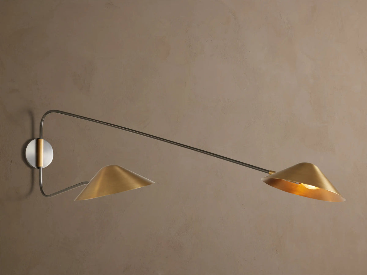 Close-up of one light from Simms Long Arm Double Sconce. It has a brass conical shade, glowing bulb, curved dark arm, and round backplate.