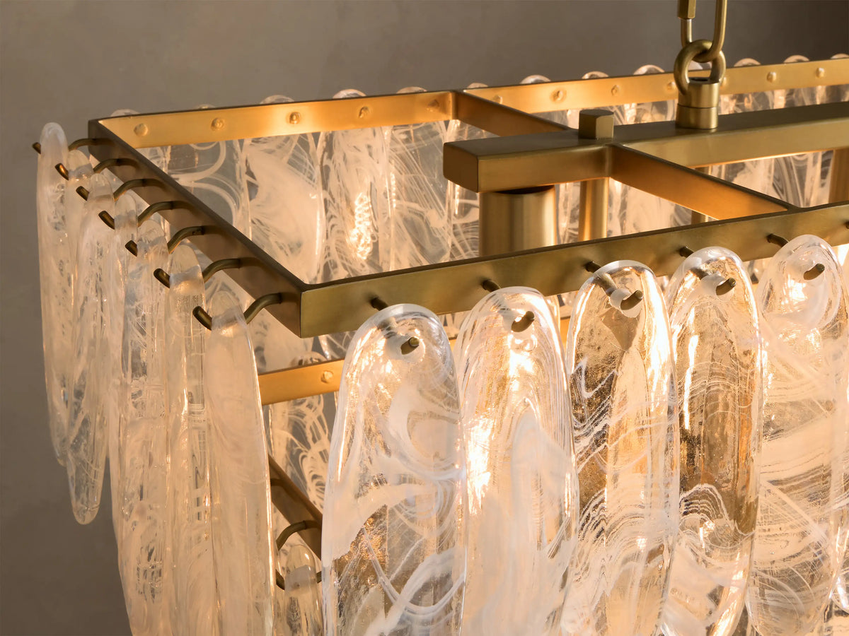 Close-up of the angled view of a rectangular brass chandelier with clear glass panels featuring white swirled patterns, casting a warm glow.