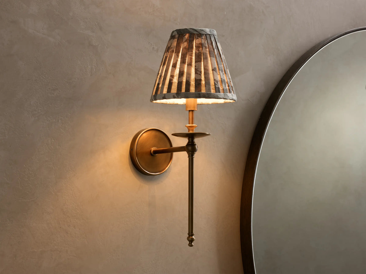 Front view of the lit antique brass Winslow Single Sconce, featuring a pleated dark patterned fabric shade, mounted on a wall.