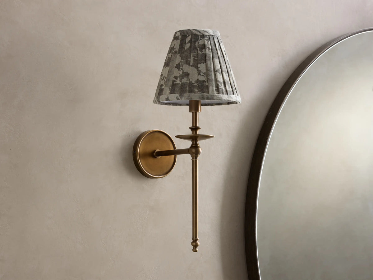 Front view of an antique brass Winslow Single Sconce with a pleated gray floral fabric shade, mounted on a light wall next to a mirror.