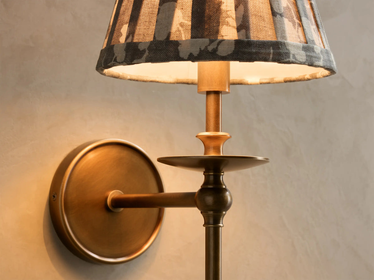 Close-up of the antique brass Winslow Single Sconce with a pleated grey and cream floral fabric shade, lit.