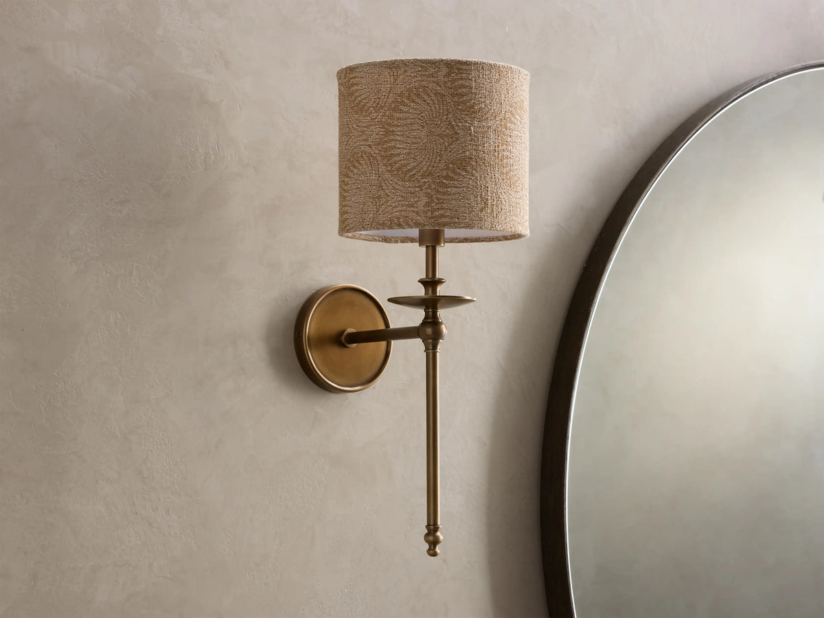 Close-up of the lit Winslow Single Sconce, showing its antique brass finish and a glowing, light brown textured fabric shade.