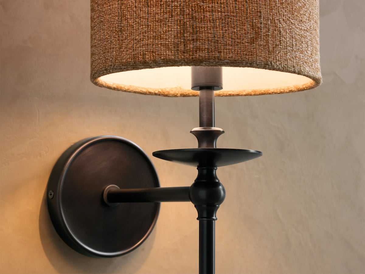 Bronze Winslow Single Sconce, illuminated, with a neutral fabric shade, mounted on a textured wall next to a round mirror.
