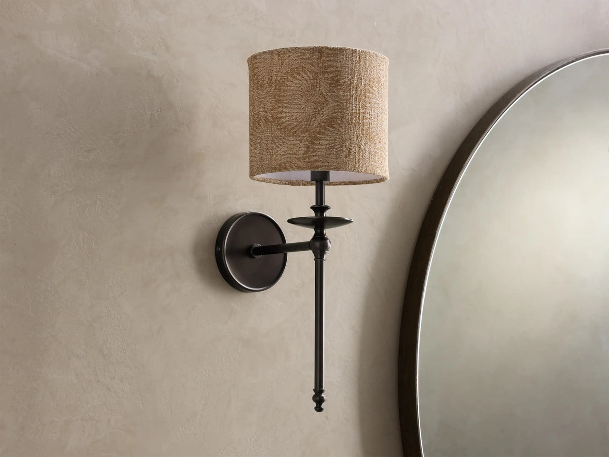Close-up of a bronze Winslow Single Sconce with a lit, textured light brown fabric shade, mounted on a light wall.