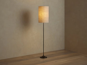 A tall, cylindrical floor lamp with a textured beige shade, emitting warm light. It has a slender black base, standing on a wooden floor.