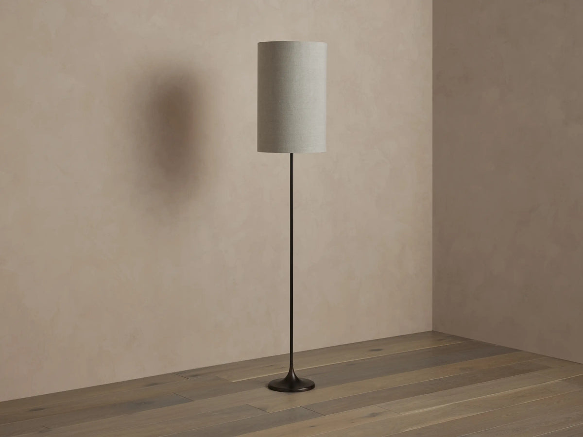 A tall, cylindrical floor lamp with a textured beige shade, emitting warm light. It has a slender black base, standing on a wooden floor.
