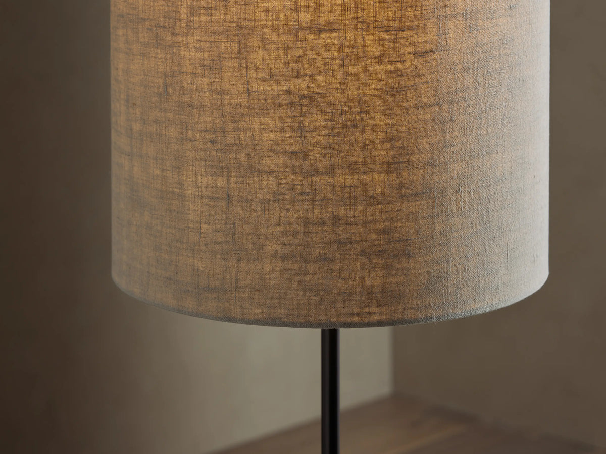 A tall, slender floor lamp with a cylindrical beige shade on a black metal stand, set against a light brown wall and wood floor.