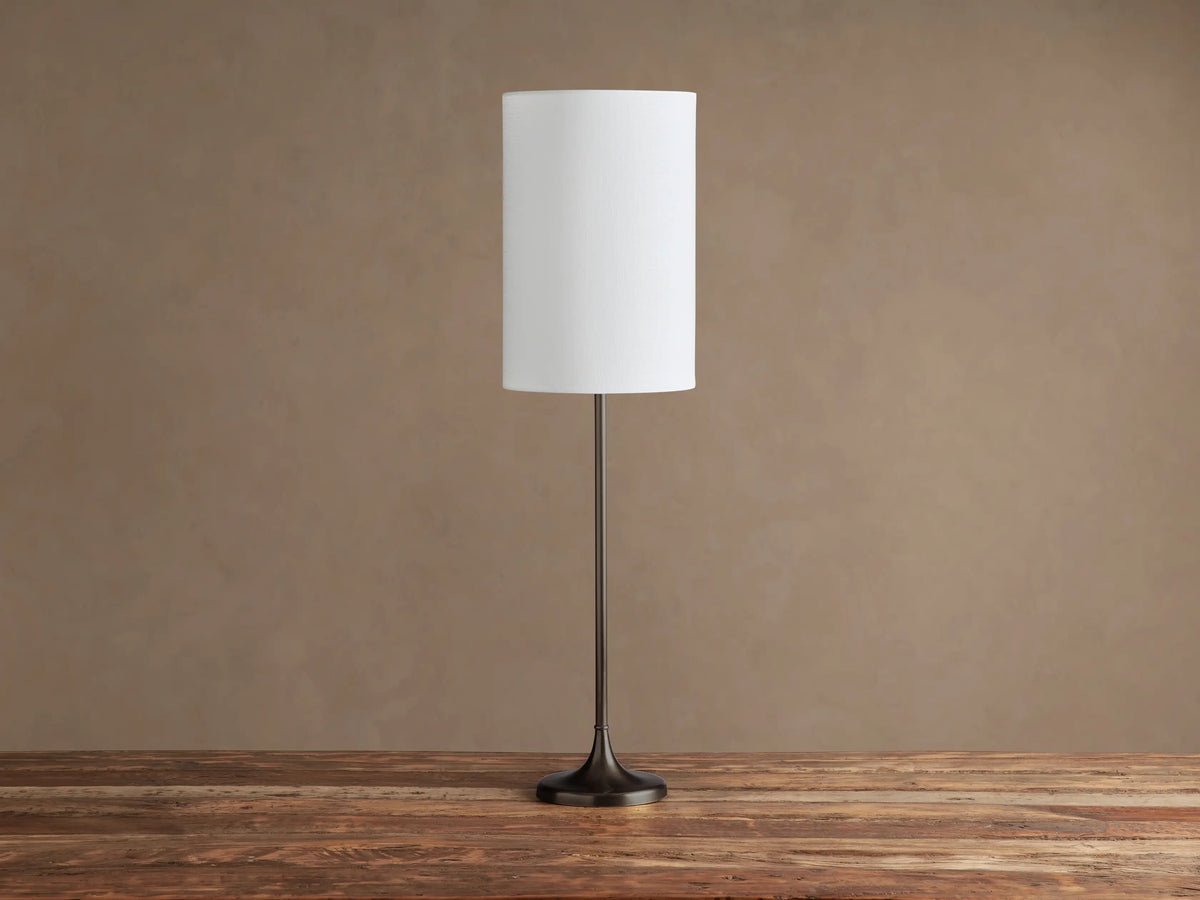 Lorelei Table Lamp: Slender dark bronze base & stem, cylindrical white linen shade, illuminated, on rustic wood.