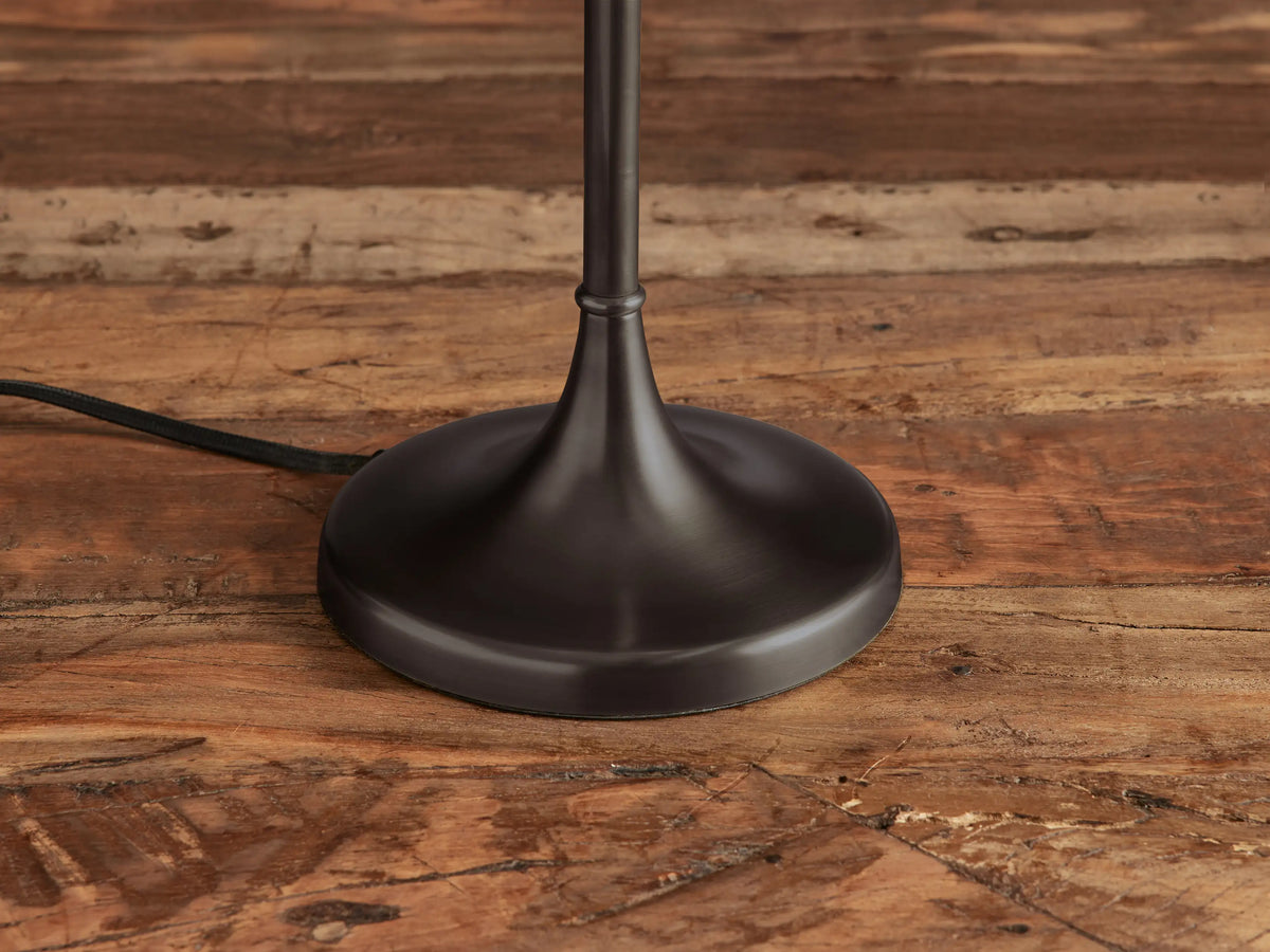 Lorelei Table Lamp with a tall white cylindrical shade, dark bronze slender stem & round base on a wooden table.