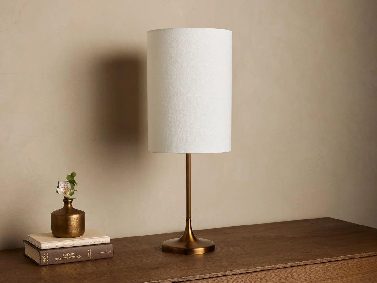 Lit Lorelei Table Lamp with a flax linen cylindrical shade and antique brass base on a wooden dresser, casting a warm glow.